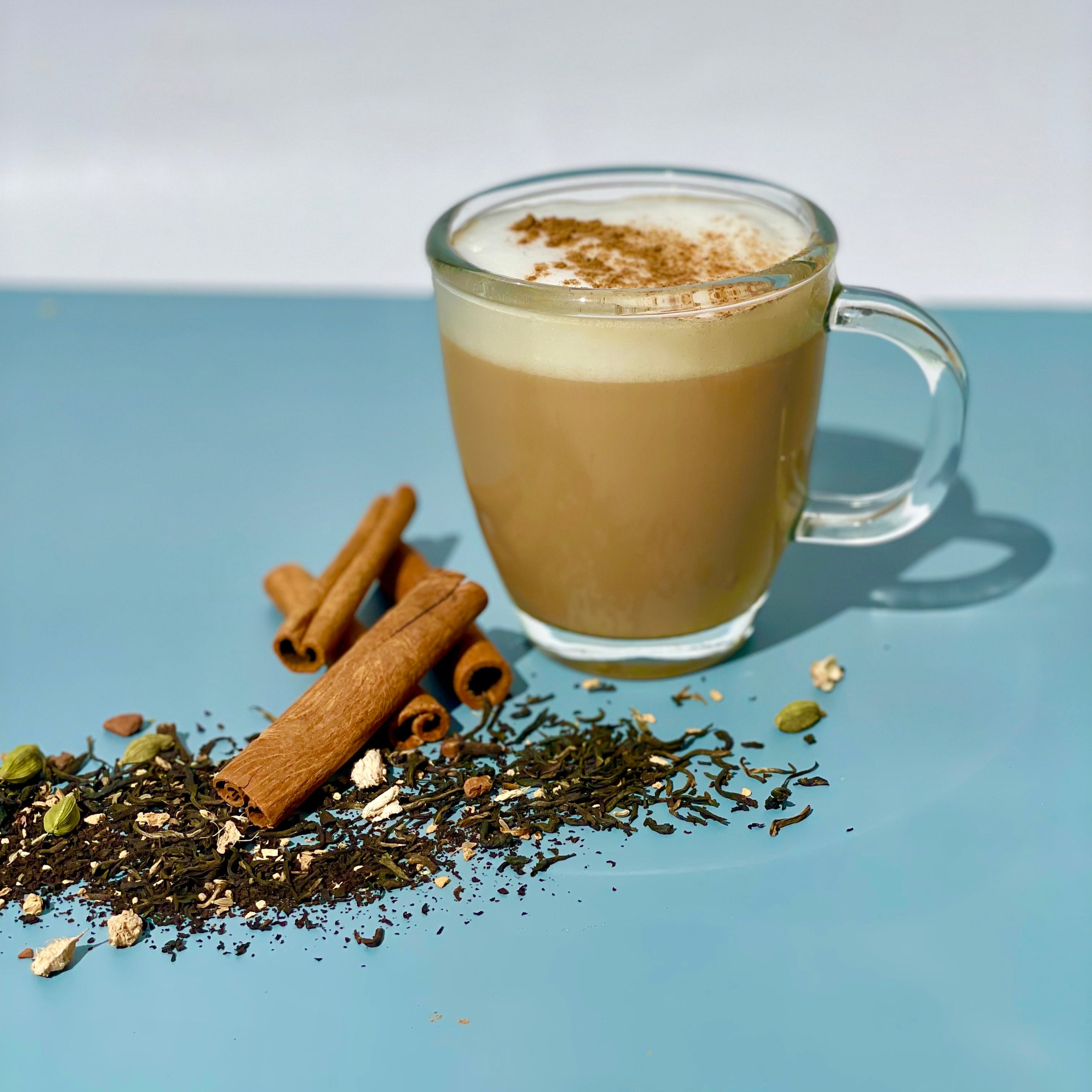 Clear glass mug with a chai latte, cinnamon sticks, and loose tea leaves on a light blue background.